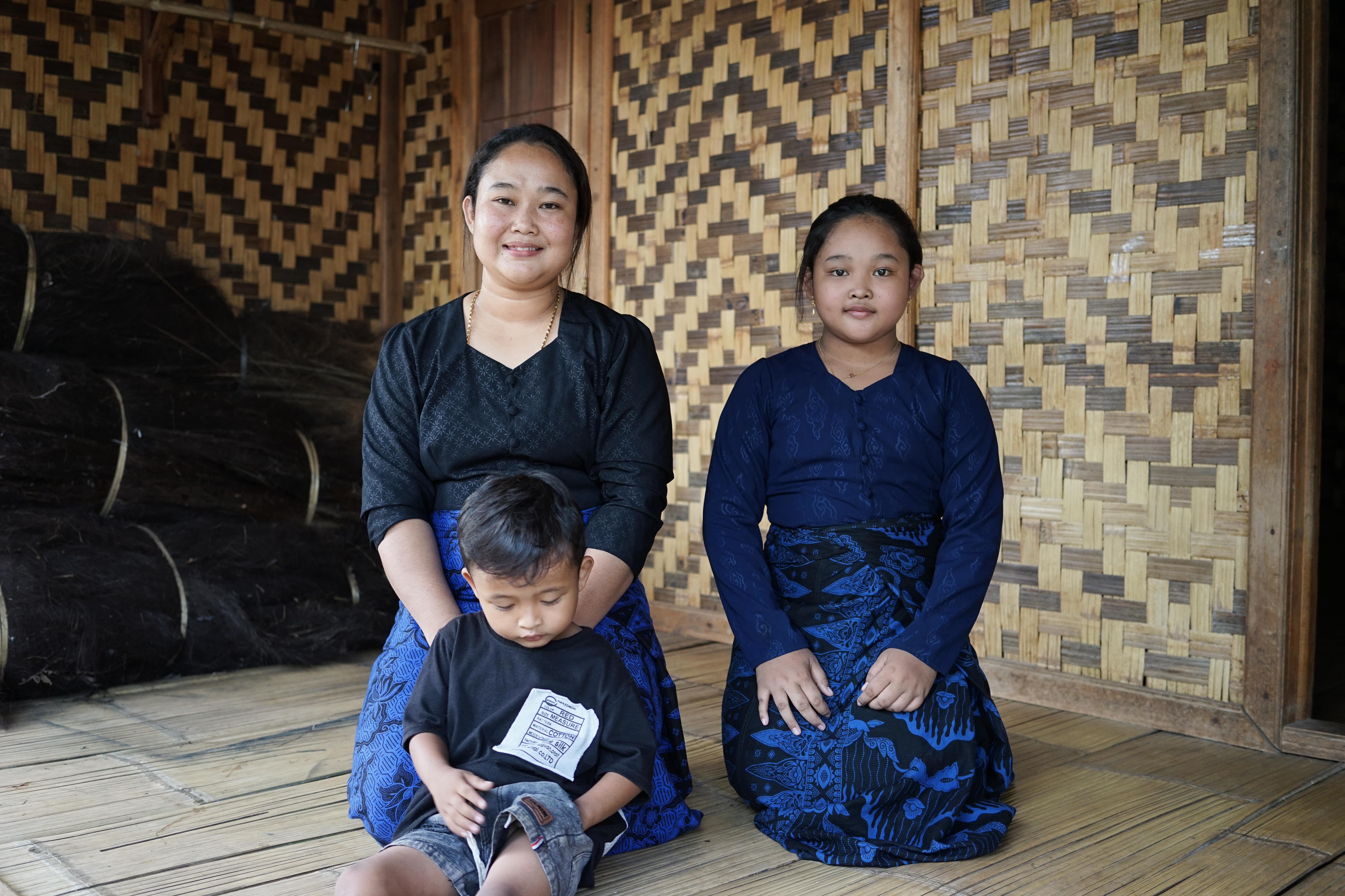 DSC06060-min A mother, her daughter, and her young son sitting on a wooden floor against a backdrop of woven bamboo walls.