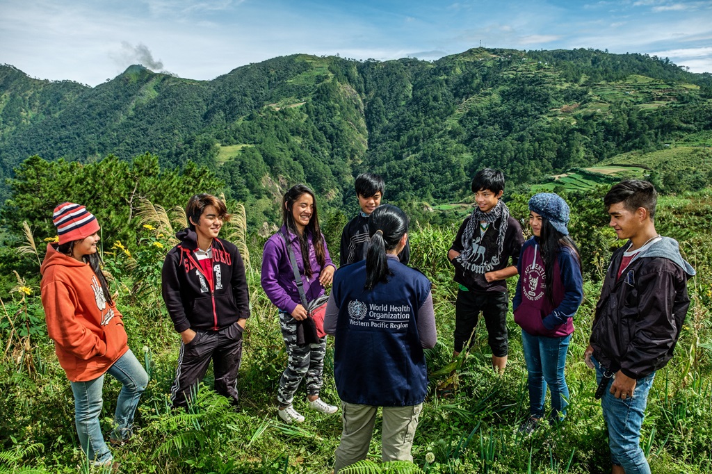 20230219_CONNECT_Savannakhet-2308 A WHO staff member stands, with their back to the camera, in the middle of a group of smiling teenagers in Benguet, Philippines. Benguet’s lush highlands can be seen in the background.